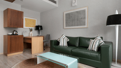 A modern room at The Atara, Gurugram featuring a green leather couch, a glass coffee table, a wooden desk with a chair, and a compact kitchenette area with a kettle