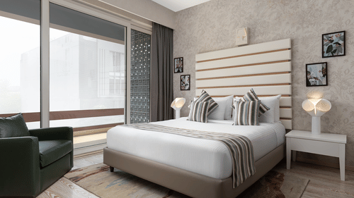 A stylish room at The Atara, Gurugram boasts a comfortable bed with a patterned runner, a plush green armchair, and a large window that offers a scenic outdoor view