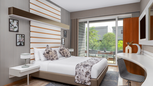A modern bedroom at The Atara, Gurugram featuring a neatly made bed with a floral runner, a stylish white desk with an armchair, and a large window offering a refreshing view of lush greenery