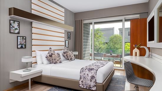 A modern bedroom at The Atara, Gurugram featuring a neatly made bed with a floral runner, a stylish white desk with an armchair, and a large window offering a refreshing view of lush greenery