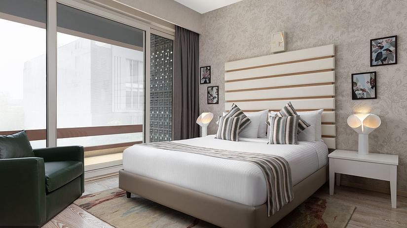 A stylish room at The Atara, Gurugram boasts a comfortable bed with a patterned runner, a plush green armchair, and a large window that offers a scenic outdoor view