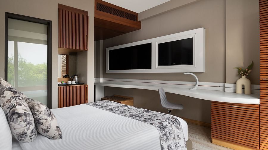 A well-appointet room at The Atara, Gutrugram featuring a functional desk area, two wall-mounted televisions, and a plush bed. The room showcases modern design and warm wooden elements