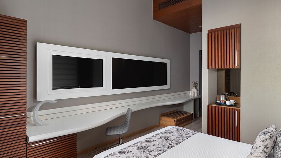 A contemporary room at The Atara, Gurugram featuring a wall-mounted TV, a sleek desk, and a bed adorned with a white and gray floral runner. The room has warm lighting and wood accents
