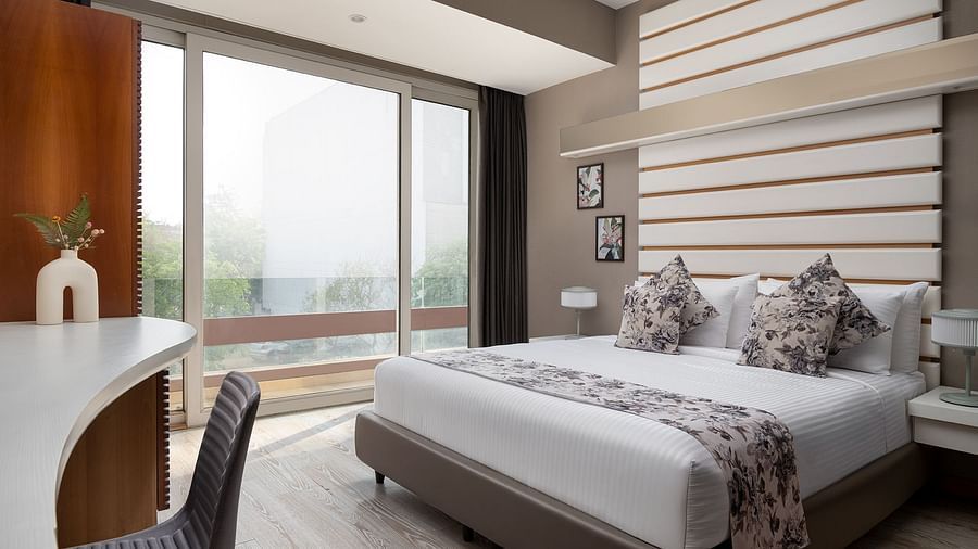 A bright room at The Atara, Gurugram boasting a large window offering an outdoor view, a comfortable bed with a floral runner, and a minimalist white desk with a chair