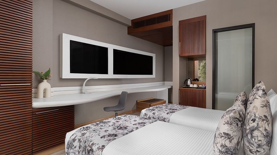Modern room at The Atara, Gurugram featuring two beds, wall-mounted TV, and a desk. The room features warm wood accents and light-coloured walls