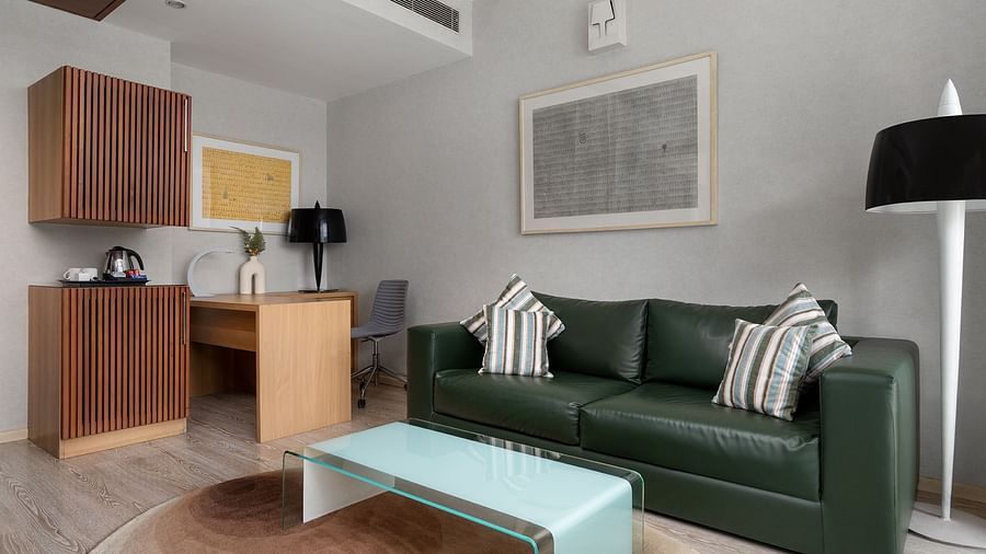 A modern room at The Atara, Gurugram featuring a green leather couch, a glass coffee table, a wooden desk with a chair, and a compact kitchenette area with a kettle