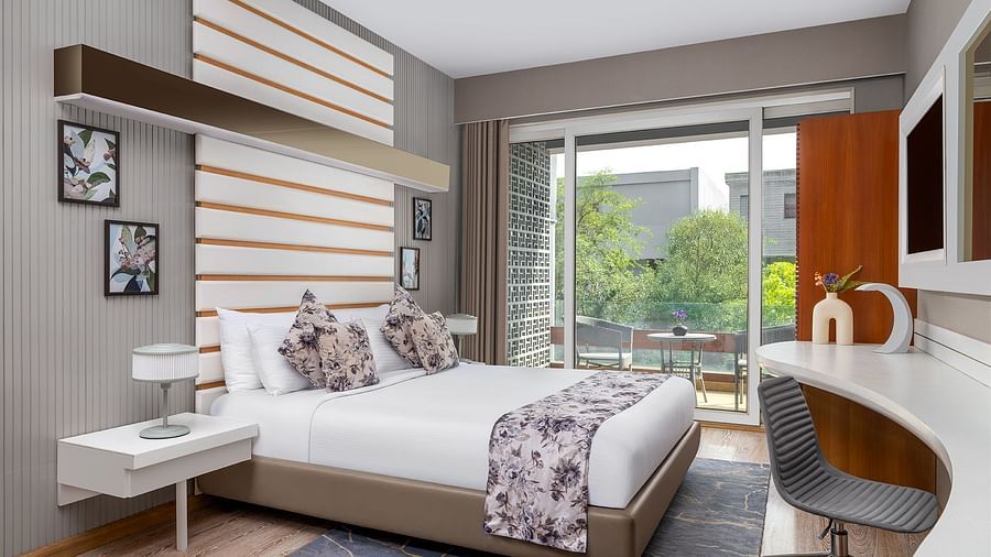 A modern bedroom at The Atara, Gurugram featuring a neatly made bed with a floral runner, a stylish white desk with an armchair, and a large window offering a refreshing view of lush greenery