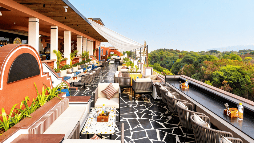 Rows of dining tables, chairs, and outdoor lounge seating line a spacious open terrace at The Sierra - By The Lake, next to a lush green forest view.