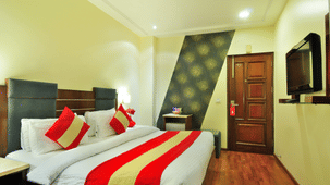 A room with a double bed featuring white linens, red and gold accent pillows, and a matching bed runner. The room includes a wooden floor, wall-mounted TV, stylish patterned accent wall, bedside table with a red lamp, and a wooden door.