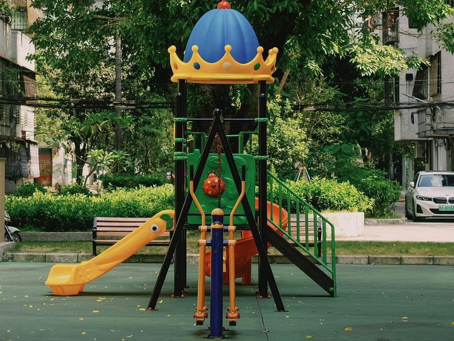 A colourful playground with a blue dome is centred in the frame, surrounded by lush green trees and some apartment buildings in the background. 