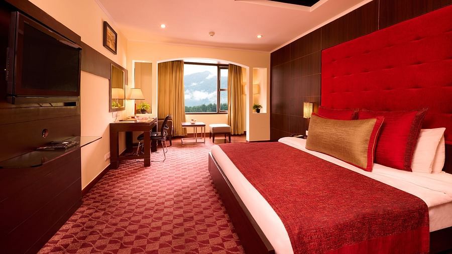 Renest Hotel Manali Warmly lit hotel room with a king-sized bed featuring a red headboard and bedding, a work desk, and a view through large windows - Renest River Country Resort, Manali