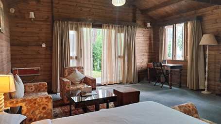 A room at The Heritage Club Tripura Castle, Shillong, with a large bed, pillows, spreads, tables, sofas, windows covered with curtains, and a lamp.