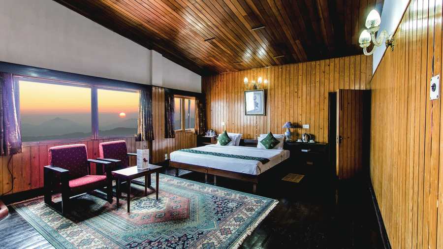 An image of a cosy room with warm wooden interiors, featuring a neatly arranged bed, sofa chairs, and a table, complemented by large glass windows offering a beautiful view of the mountains and the sun - Central Gleneagles Heritage Resort, The Mall Road, D