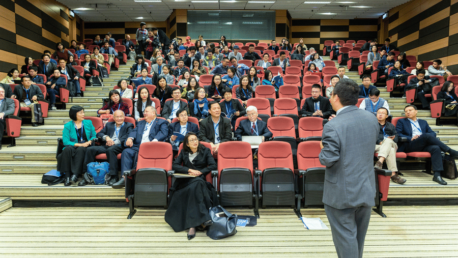 View from the stage of a speaker in a grey suit facing a diverse audience seated in a tiered lecture hall.