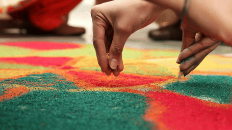 Hands sprinkling coloured powders onto a vibrant floor rangoli with red, green, yellow and orange geometric patterns during a festival.