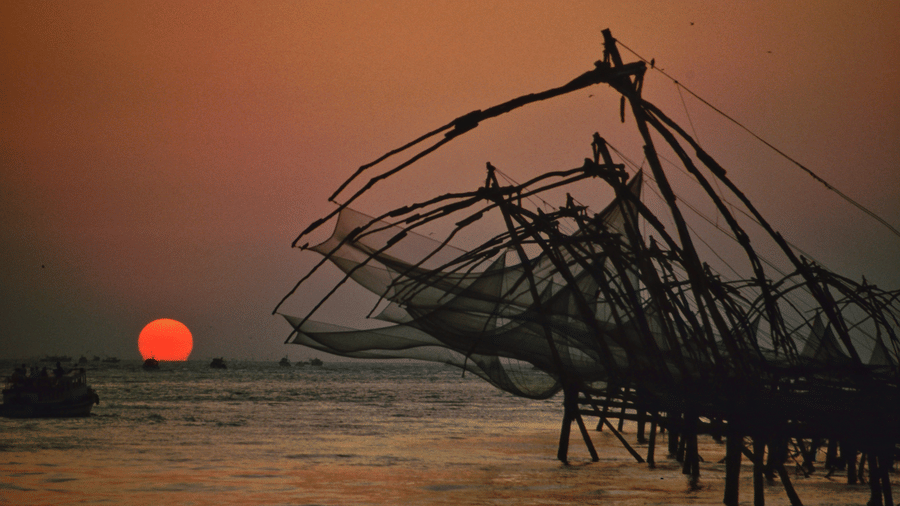 Silhouetted Chinese fishing nets and boats against a glowing orange sunset in Kochi