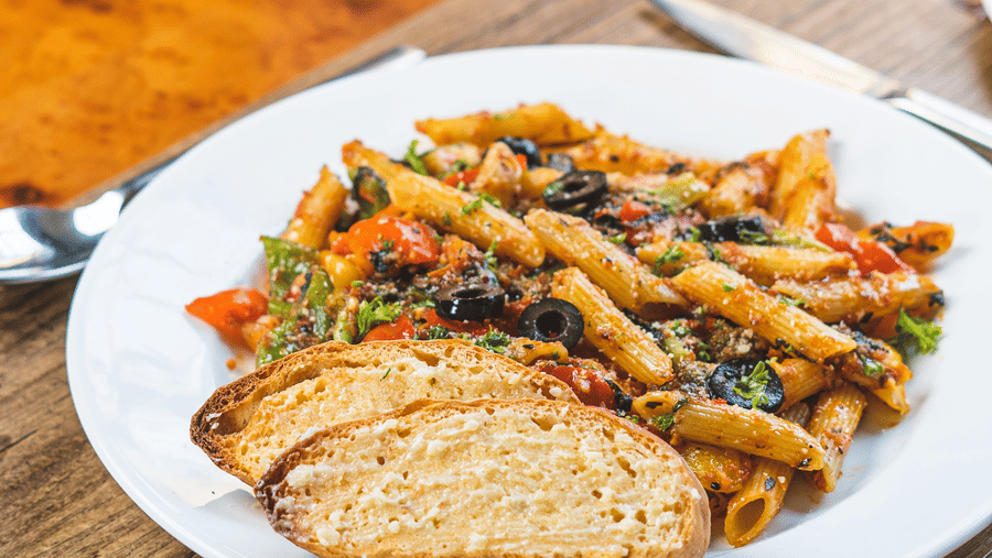 White plate of colourful pasta with vegetables and dark olives, served with 2 slices of toasted bread on a wooden table at Hotel Hukam's Lalit Mahal.