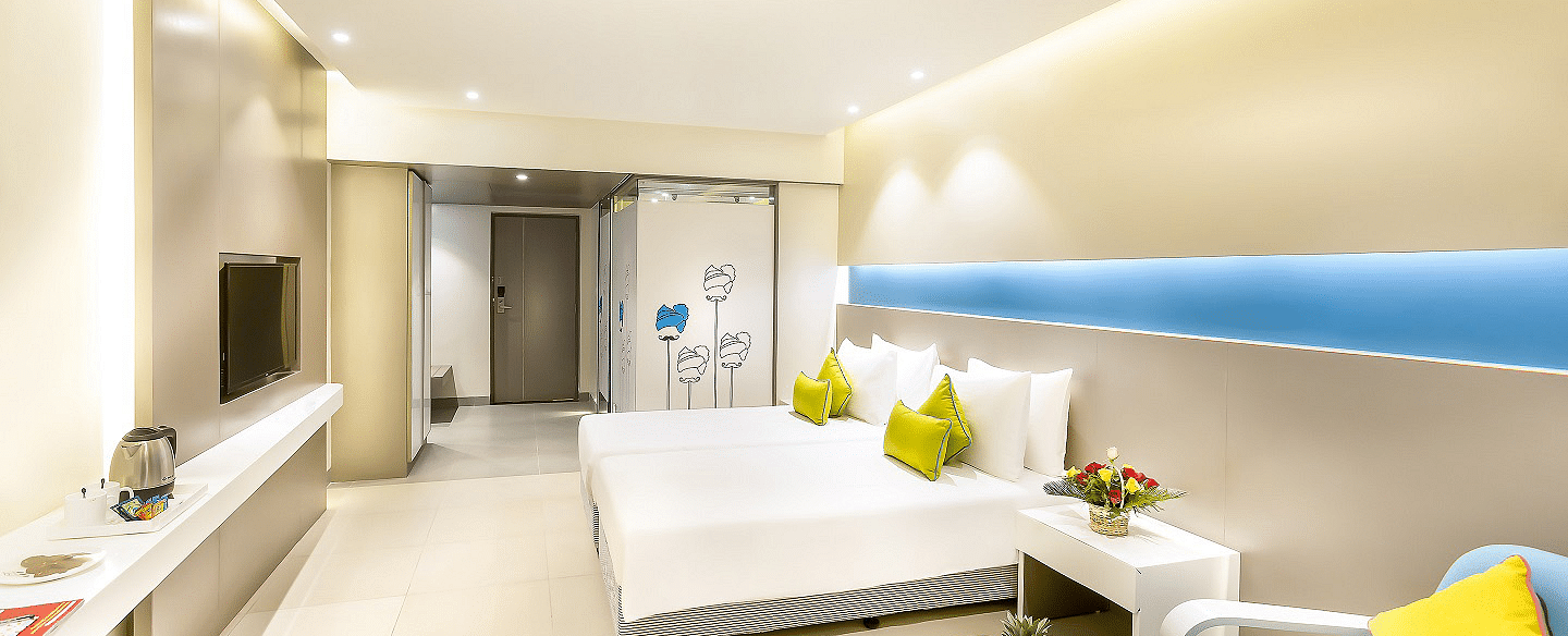 A spacious room features a large bed with a headboard stretching across the wall. Cushion chairs sit beside a wooden bedside table, and a wall-mounted television is positioned opposite the bed