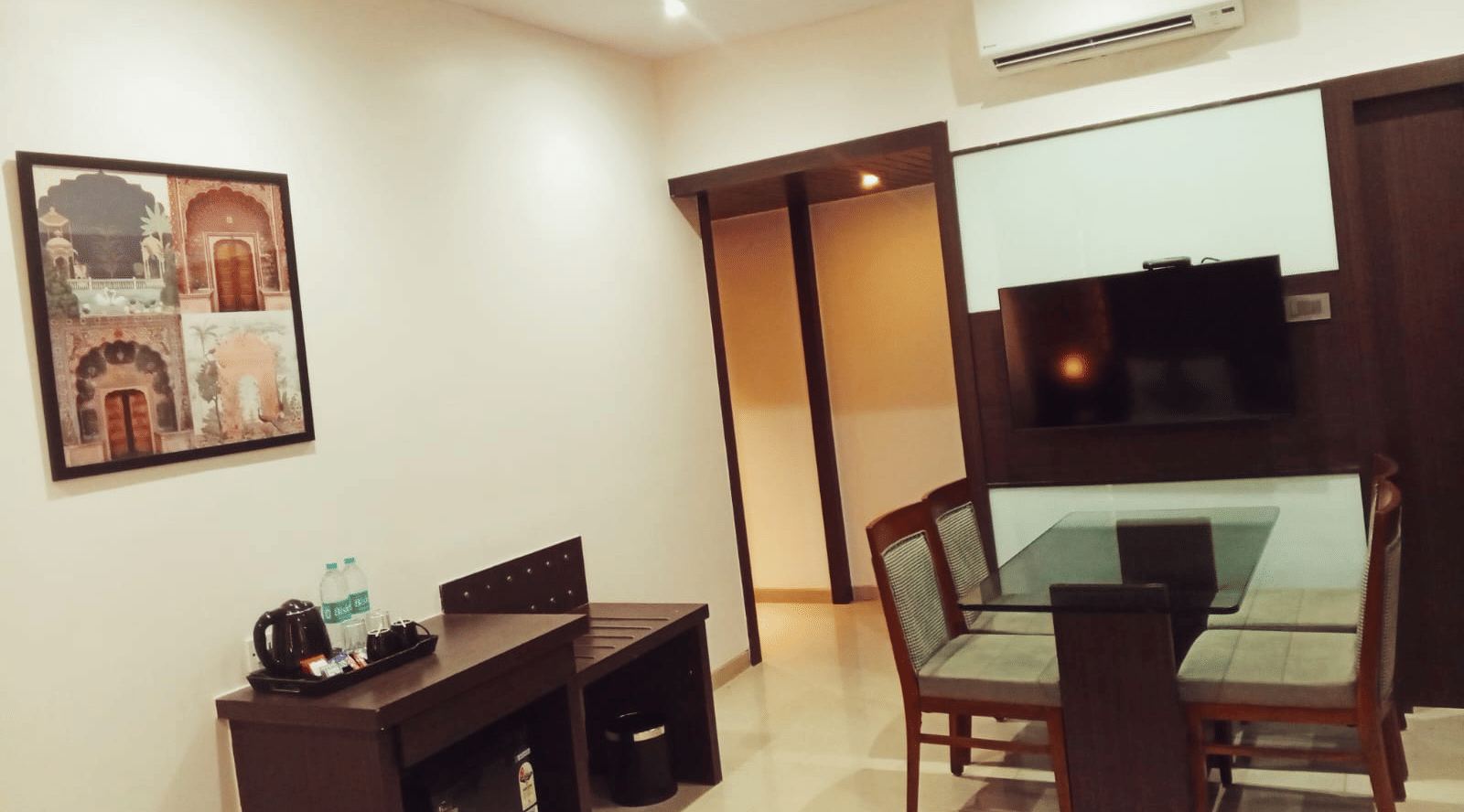 A bedroom at Hotel Mumbai House, Thane featuring a dining area with a glass top table, chairs, a dark refreshment table with a kettle, a framed painting, and a television.
