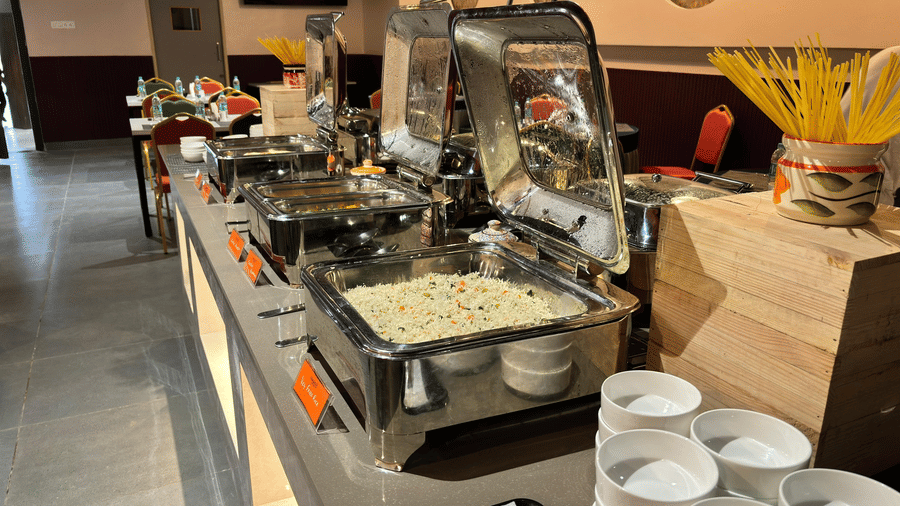 A professional buffet counter in a hotel dining room, featuring stainless steel chafing dishes and a selection of fresh food and garnishes at Apartel by Aarin, Oragadam.