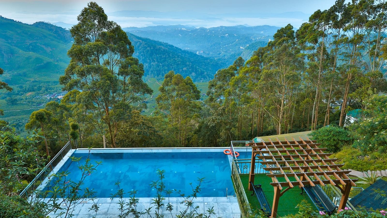 Aerial view of a swimming pool nestled in a lush green forest, with mountain ranges in the background – Fragrant Nature Munnar.
