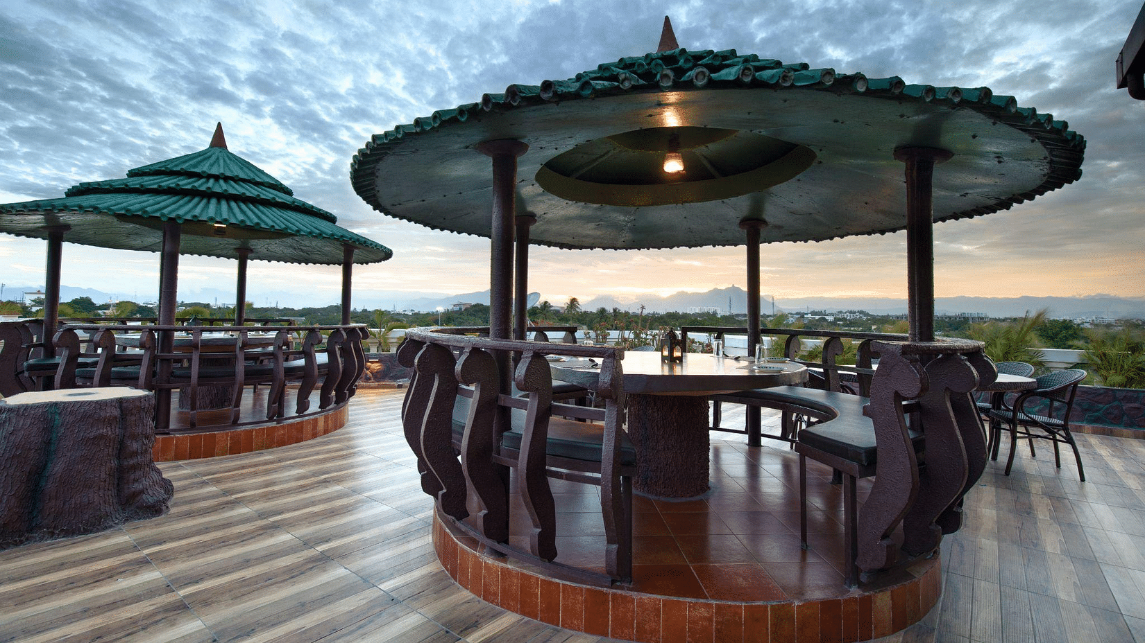Outdoor dining area with several covered gazebos featuring tables and chairs on a wooden deck, set against a dramatic, cloudy sky at Jenneys Residency, Coimbatore