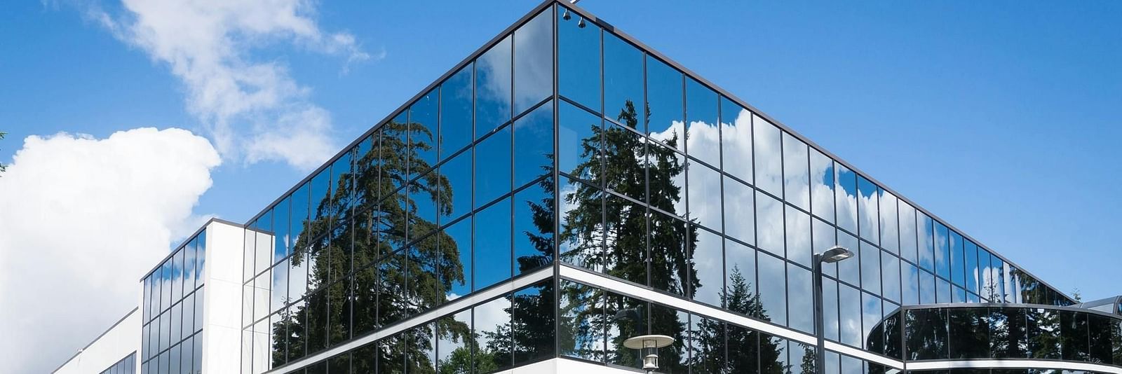 A modern, angular office building with glass walls reflecting the blue sky and surrounding greenery.