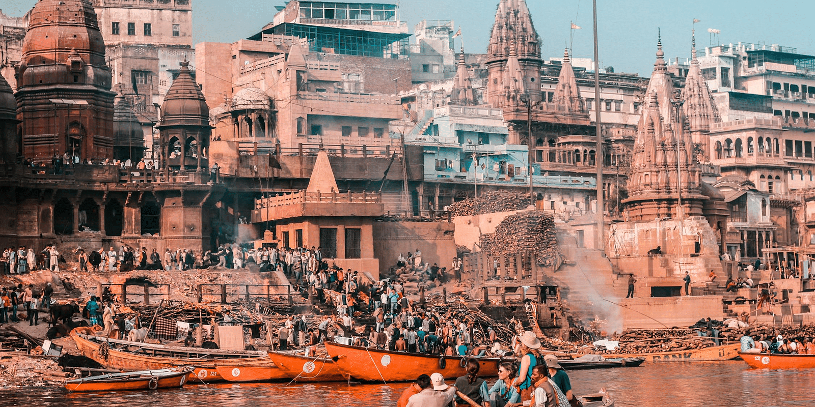 An image featuring a few people in boats on the river, with the ghats of Varanasi and buildings in the background.