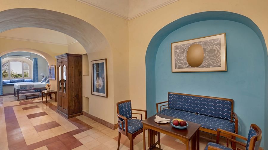 Hotel hallway with arched openings, a seating area with a blue sofa and chairs around a table, artwork, and a view into a bedroom at Tijara Fort-Palace, Alwar.