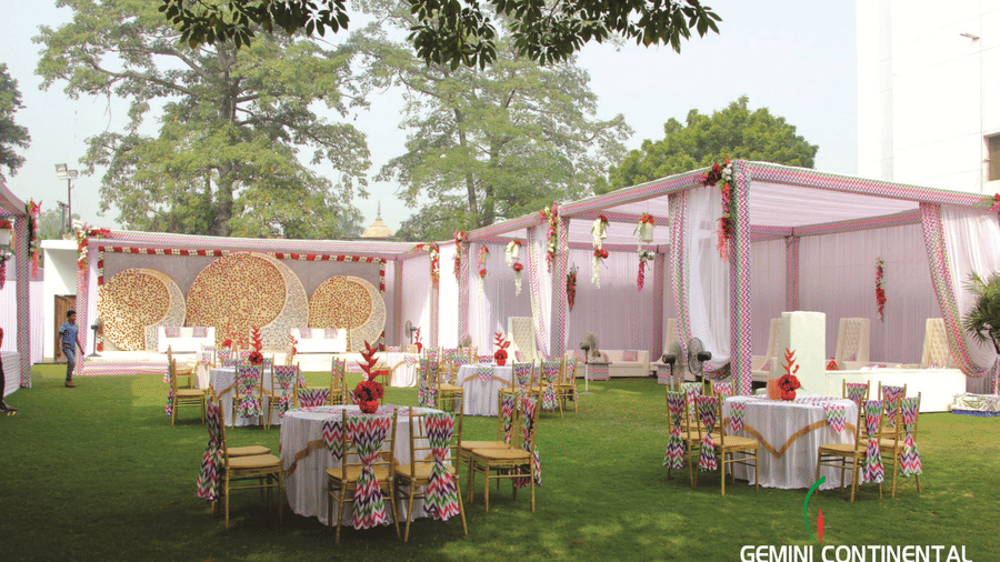 Outdoor lawn venue with round tables and chairs set beneath tall trees, a tented canopy in the background and open daylight overhead.