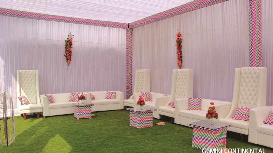 Outdoor lawn lounge area with white tufted seating, patterned cushions, hanging floral decor overhead and a tented canopy above the whole space.