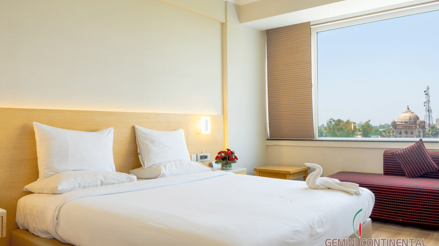 Hotel room with a bed dressed in white linen, a maroon sofa placed nearby, a large window with a city view and light-toned interiors all around.