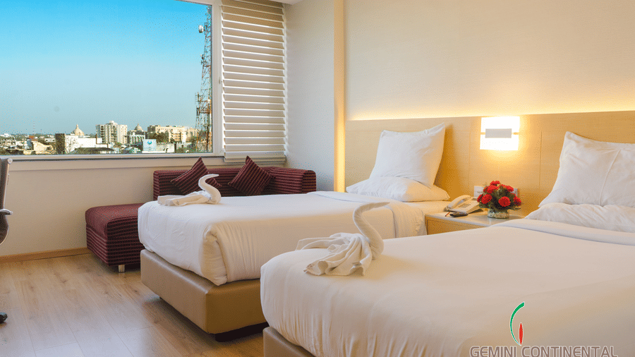 Hotel room with twin beds dressed in white linen, maroon footstools, a large window with a city view and a fruit basket on the bedside table.