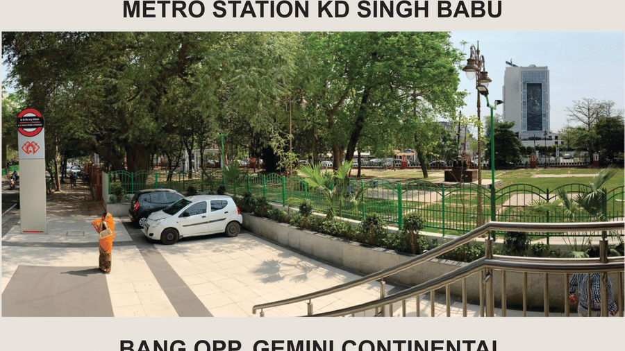 Street view with a metro station signboard, a landscaped park with pathways, parked vehicles and city buildings visible in the background nearby.