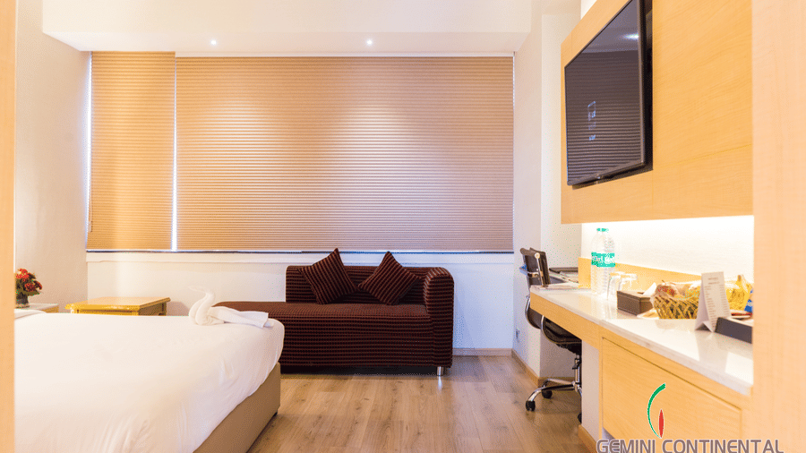 Hotel room with a bed dressed in white linen, a maroon sofa, a wall-mounted television, a wooden work desk and light wooden flooring throughout.