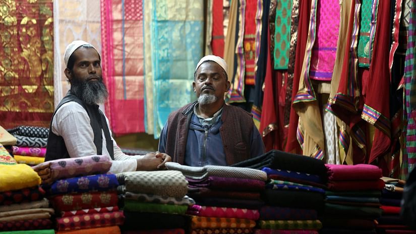 Two men sitting among stacks of vibrant, folded fabrics in a colorful textile shop, surrounded by richly patterned sarees hanging in the background.