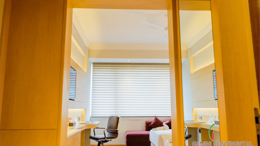 Hotel room entrance area with a wooden sliding partition, a work desk and chair, window blinds and a bed visible in the background nearby.