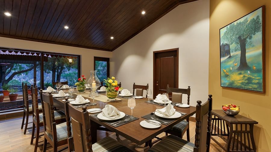 An image of a  large wooden dining table with wooden chairs and crockery for 8 people, pictured with a painting that is on the wall.