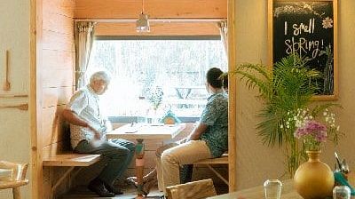 Indoor dining table at Karma Chalets with food and drinks set up, and a person seated near a sunlit window.