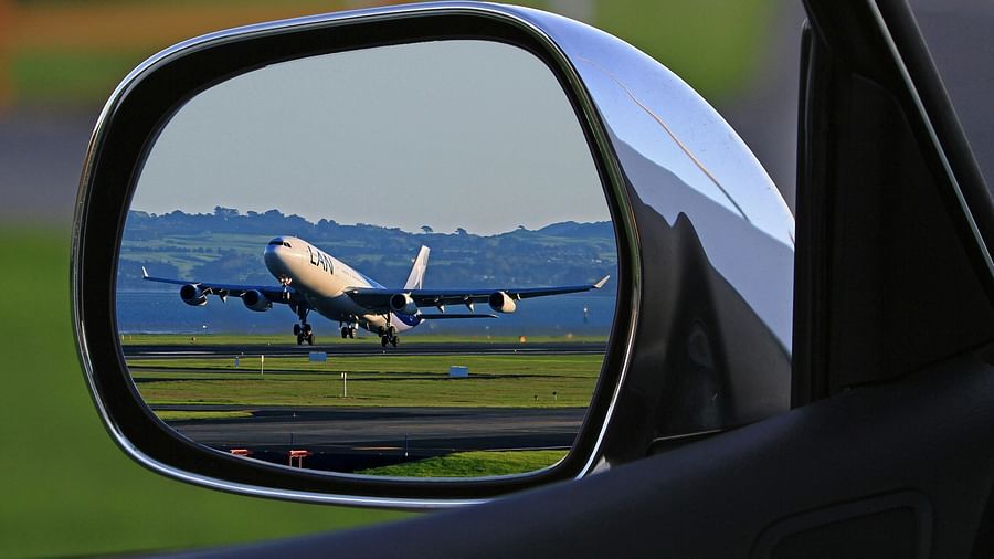 An aeroplane ascends from a runway, its image reflected in a car's side mirror, showing the aircraft against a landscape.