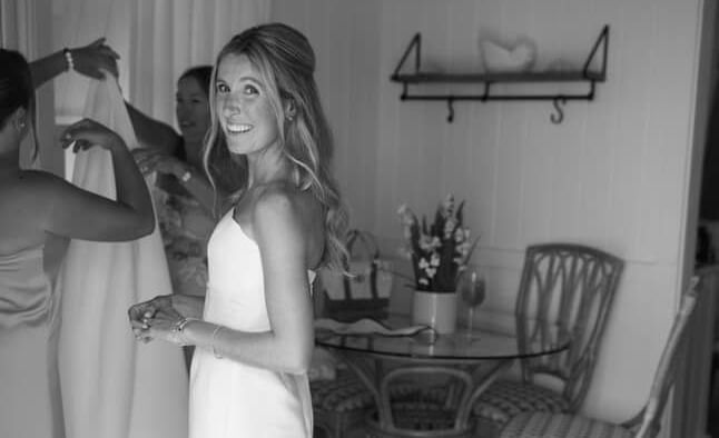 Person in a wedding dress stands indoors holding a bouquet, with another person adjusting the dress, chair and wall art in the background at Abaco Inn.