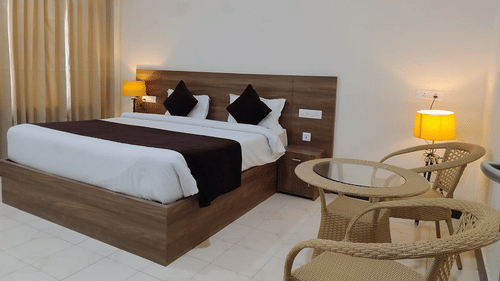 Deluxe Room at Pride Resort Rudraprayag featuring a queen size bed with wooden headboard and lamps on both sides and sitting space showcasing a pair of chairs and a coffee table next to it