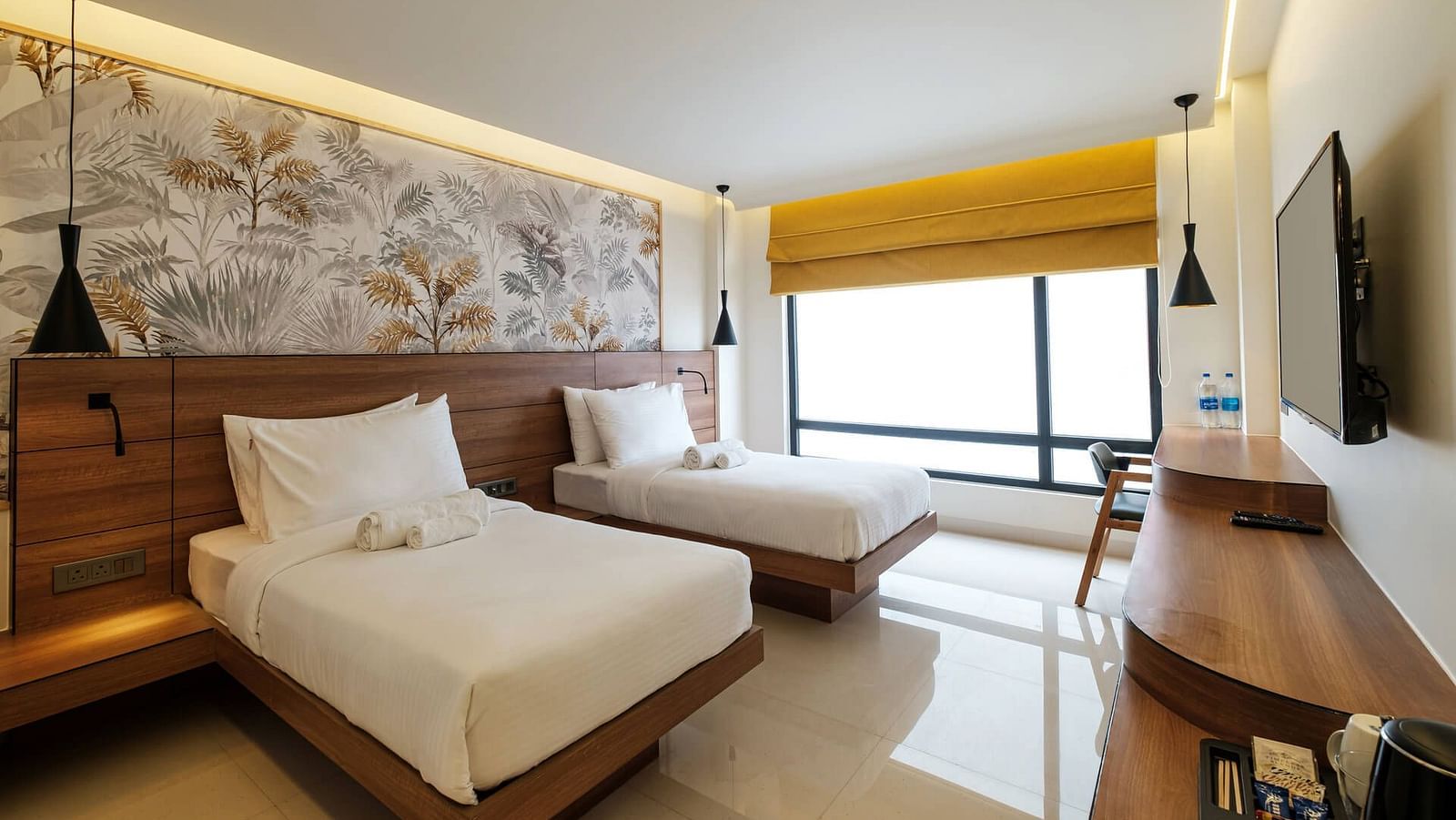 A spacious room at Indie Stays, Jaipur, with two beds and a joined wooden headboard, wall-mounted TV, wooden desk with a chair, and large alcove windows.