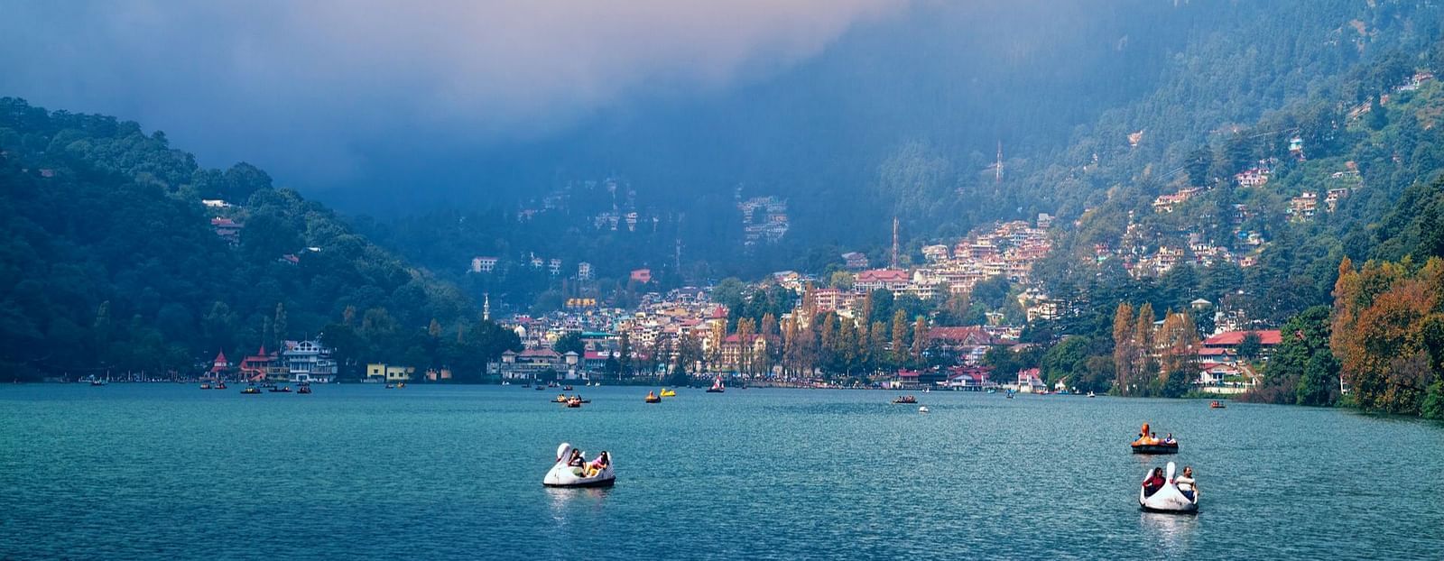 A serene view of Nainital Lake with boats floating on calm waters, backed by colourful hillside houses under a soft, cloudy sky.