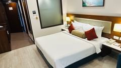 Contemporary hotel room with a neatly made double bed, accent pillows, bedside tables with lamps, wall art above the headboard, and a hallway leading to the entrance - Sobit Sarovar Portico, Goa.