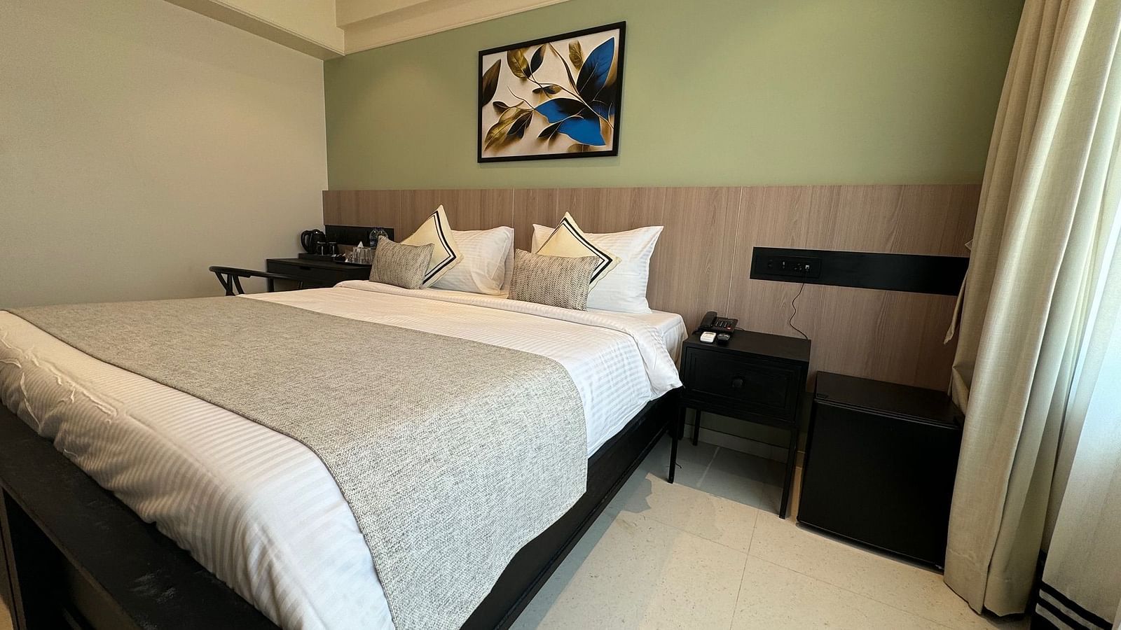 Side view of the king bed with a painting hung above and a side table next to it inside deluxe room - Hotel Mumbai House, Valnai, Malad