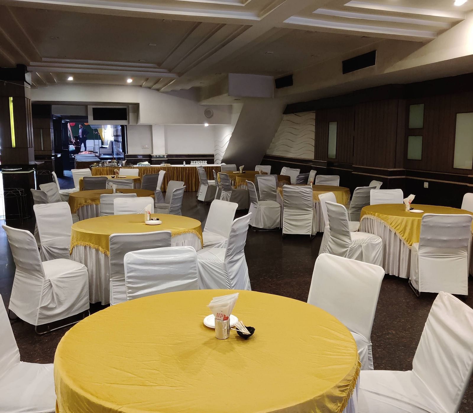 A few round tables covered with bright yellow coloured table cloth and chairs surrounding it with a stage at the far end of the Banquet Hall at Hotel Gold, Panipat.