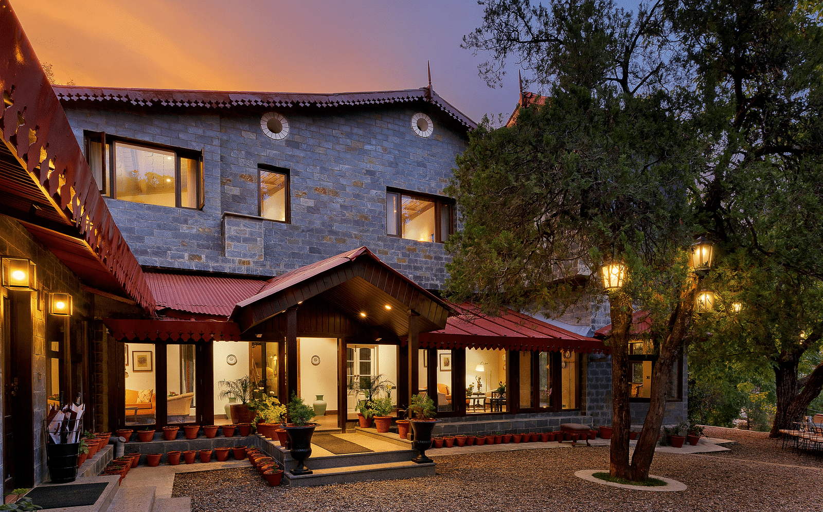 An Image of facade of  building with a lot of wooden details, which stands behind a Twin tree with lights, captured in the photo after the golden hour - The Naini Retreat