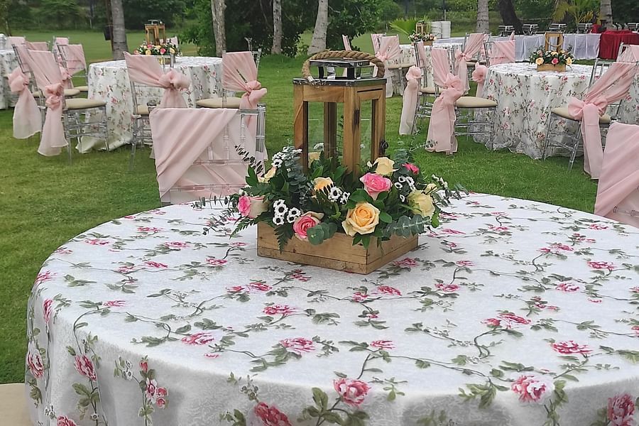 Round outdoor dining table with a floral tablecloth, topped with a wooden lantern and flower arrangements, set on a green lawn with other tables in the background - Black Thunder, Coimbatore