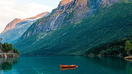 A calm lake with a single orange boat floating in the water, surrounded by green hills and tall rocky mountains with patches of snow on top.